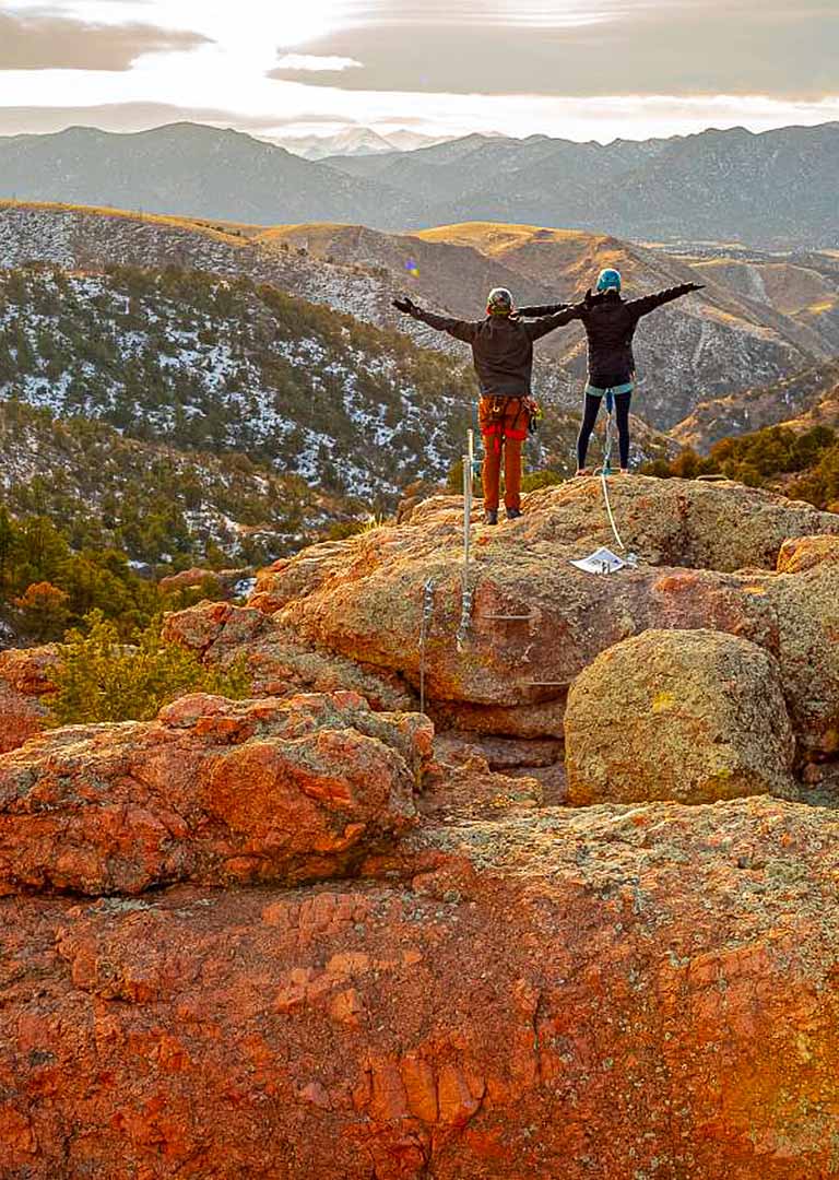 Two people on a rock outcrop above the canyon with the mountains behind